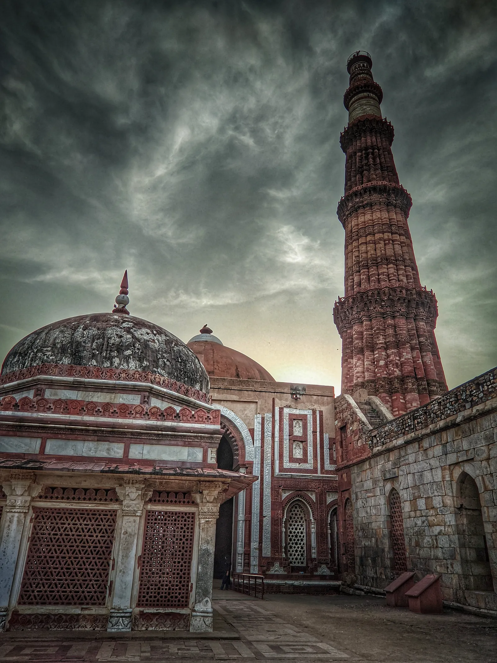 A tall fluted brick tower beside ruined domes under a dark sky