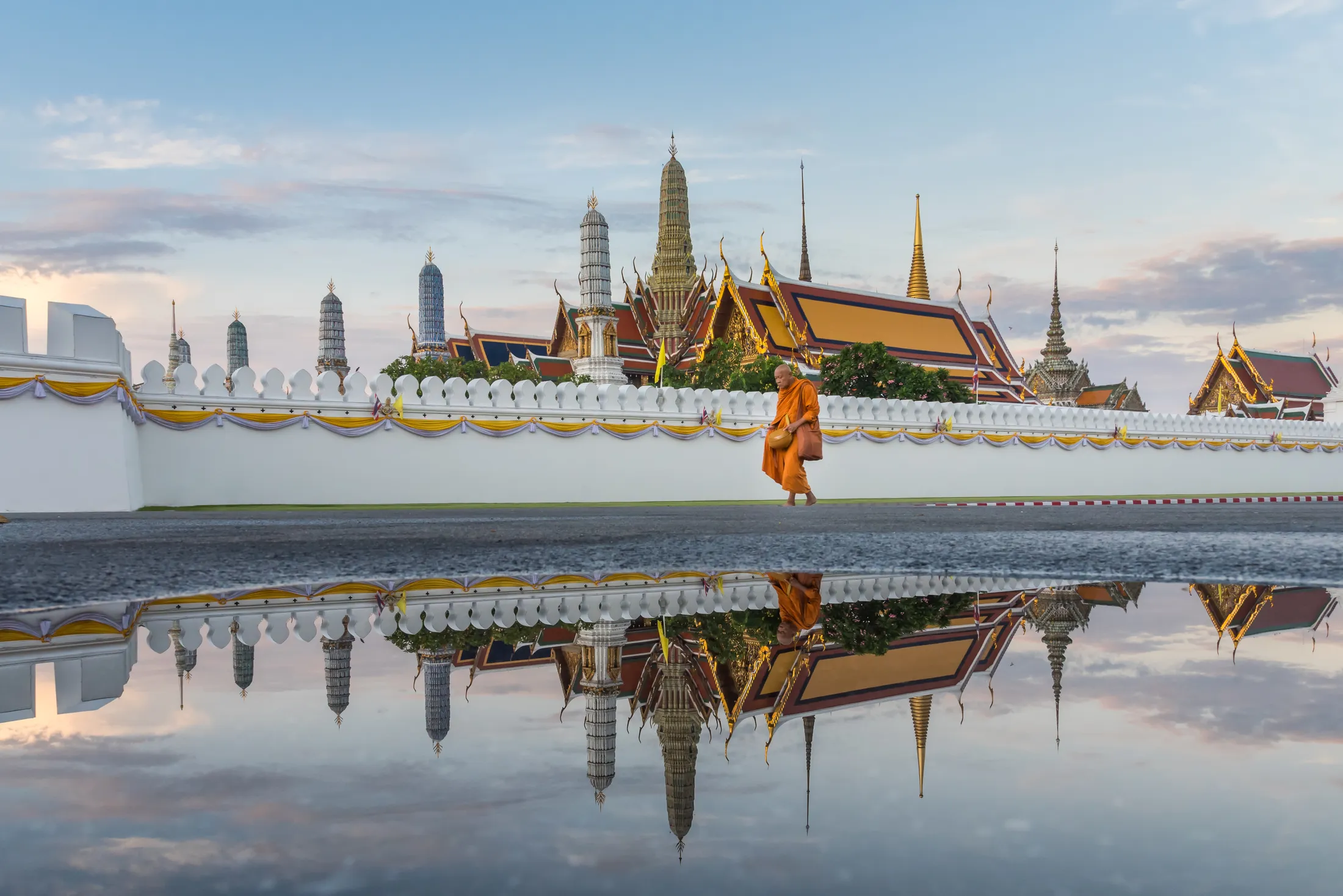 The spires of Wat Phra Kaew over the palace wall, a monk walking past, mirrored in standing water